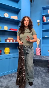 Γυναίκα με σγουρά μαλλιά, φορώντας γκρι πουκάμισο και τζιν, κρατά ένα καφέ υφάσμα με πουά σε ένα μπλε δωμάτιο με ράφια και διακοσμητικά αντικείμενα.