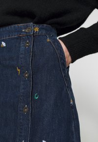 Denim skirt featuring a dark blue hue, front button closure, side pockets, and colorful embroidered details including suns and clouds.