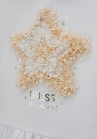Tessuto bianco con un design a stelle testurizzato realizzato con petali color crema e beige, con testo metallico argento che recita "GUESS festeggiamo!"