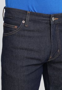 Tmavě modré džíny z denimu se střihy rovného nohavice, které mají viditelné švy, embosovaný knoflík a měděné akcenty na kapsách.