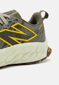 Gros plan d'une basket en mesh vert olive avec logo jaune, semelle blanche épaisse et boucle au talon, montrant une partie de la semelle sur un fond clair.
