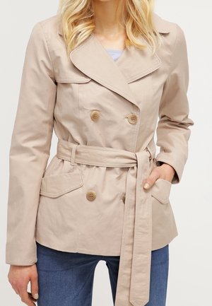 Beige double-breasted belted jacket with wide lapels, buttoned pockets, worn by a person with one hand in a pocket and blue jeans.