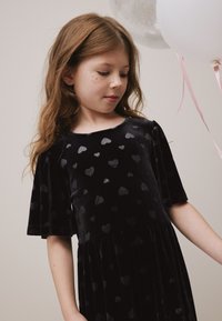 Vestito in velluto nero con motivo a cuori, maniche corte, scollo rotondo e gonna a balze. La texture morbida ne accentua l'aspetto visivo.