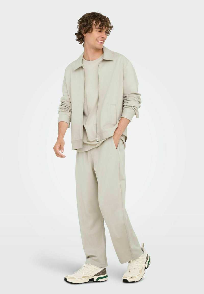 Veste zippée gris clair avec poches latérales, assortie d'un t-shirt à col rond et d'un pantalon ample. Baskets décontractées épaisses en crème et vert.