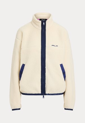 Kvinde iført en cremefarvet fleecejakke over en marineblå rullekrave og plisseret nederdel, med hvide sokker og hvide sneakers, stående foran en neutral baggrund.