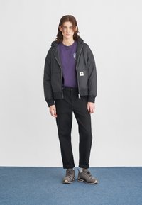 Schwarze Steppjacke mit Kapuze, zwei Vorderaschen, kombiniert mit einem lila T-Shirt, schwarzen Hosen und grauen Wanderschuhen mit schwarzen Akzenten.