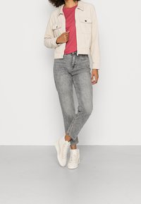 Femme portant une veste en velours côtelé beige, un t-shirt rouge, un jean taille haute gris et des baskets montantes blanches, debout devant un fond blanc uni.