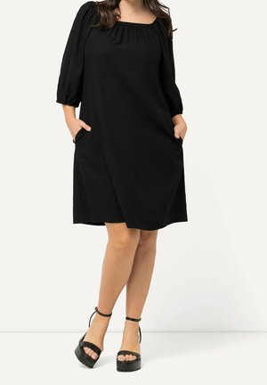 Kobieta ubrana w czarną sukienkę do kolan z trzy czwarte rękawami z bufkami i kwadratowym dekoltem, stoi z rękami w kieszeniach, ma na nogach czarne sandały na platformie.