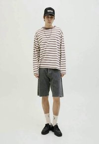 Young man wearing a striped long-sleeve shirt, black cap, gray denim shorts, white socks, and black shoes standing against a plain background.
