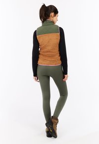 Gequilted groene en bruine fleece vest met roze afwerking, gecombineerd met olijfgroene leggings en bruine wandelschoenen, met een focus op gelaagde texturen.
