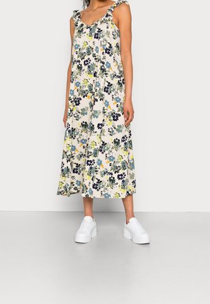 Vrouw draagt een mouwloze, gerimpelde midi-jurk met bloemenprint en witte plateau sneakers, staat tegen een effen lichte achtergrond.