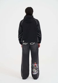 Giacca nera con cappuccio e maniche texturizzate, rifinita con piping bianco, abbinata a jeans larghi in denim scuro con un vivace design ricamato.