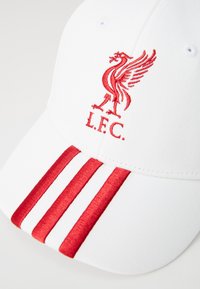 Hvid baseballcap med rødt Liverpool FC-logo og tre vertikale røde striber på skyggen mod en ensfarvet baggrund.