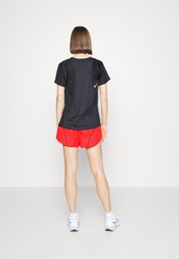 Camiseta deportiva negra de manga corta con un logo blanco, combinada con pantalones cortos rojos con costuras oscuras. Lleva puestas unas zapatillas de correr blancas.