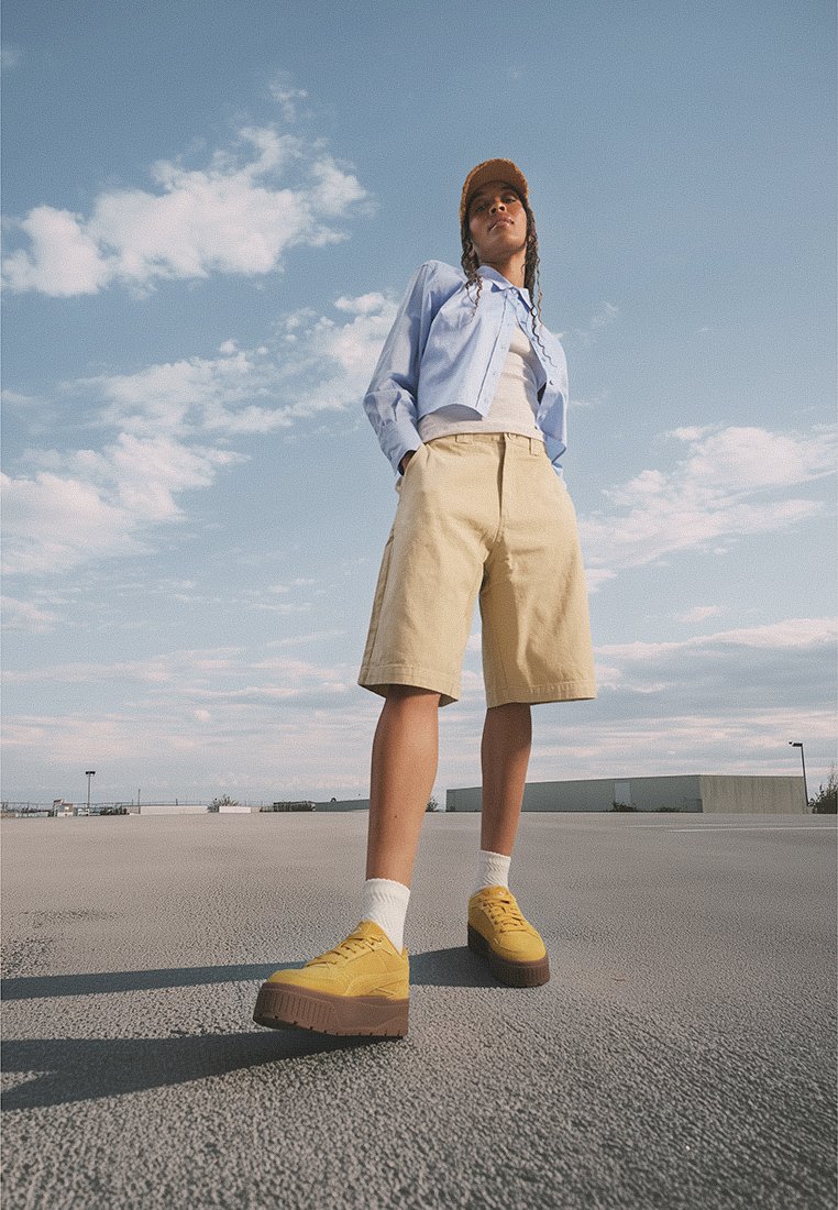 Camisa de botones azul claro, pantalones cortos beige y zapatillas de plataforma amarillas con una suela texturizada. Medias blancas visibles, al aire libre con cielo azul.