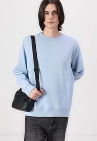 Lichtblauwe sweatshirt met lange mouwen en geribbelde cuffs, gecombineerd met een kleine zwarte crossbody tas met ritssluiting en verstelbare schouderband.