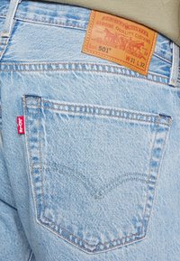 Ljust blå denimjeans med en brun läderlapp på midjan. Har en signatur röd flik på bakfickan och synliga sömmar.