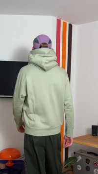 Sudadera con capucha de color verde claro con dobladillo acanalado, combinada con pantalones verde oscuro. El fondo muestra rayas coloridas en la pared y mobiliario.