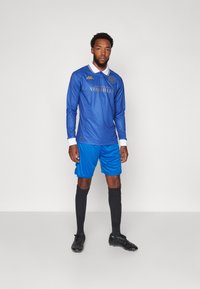 Modrá futbalová dres s dlhým rukávom a bielym golierom, so zlatým textom, doplnený modrými šortkami a čiernymi futbalovými kopačkami. Obsahuje logo Kappa.