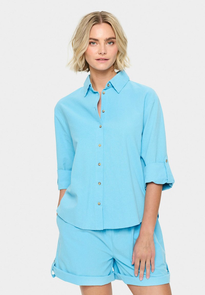 Camicia button-up azzurro chiaro con maniche arrotolate e bottoni dorati, abbinata a pantaloni corti blu coordinati. Tessuto morbido con una vestibilità comoda.