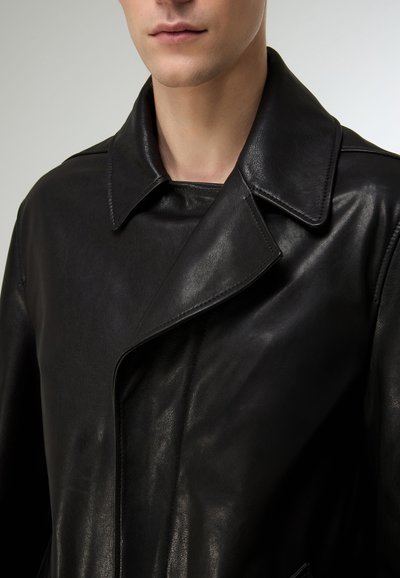 Schwarzer Ledermantel mit klassischem Kragen, glatter Textur und figurbetonter Silhouette. Auffällige Details sind die Nähte entlang der Kanten.