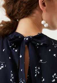 Top floreale blu navy con fiocco sulla schiena, caratterizzato da piccoli motivi floreali bianchi e un tessuto liscio e leggero.
