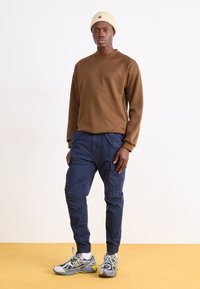Brun langermet genser med v-hals; marineblå cargo bukser med sidelommer; grå og gule joggesko; beige strikket lue.