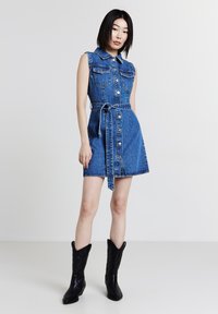 Robe en denim avec un corsage ajusté, un design sans manches, un devant à boutons et deux poches poitrine. Comprend une ceinture à nouer et une jupe évasée.