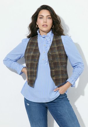 Frau mit dunklem Haar, die goldene Creolen, eine blau gestreifte Rüschenbluse, eine braune karierte Weste und blaue Jeans trägt und mit den Händen in der Hüfte posiert.