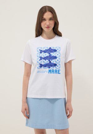 Femme portant un t-shirt blanc à manches courtes avec trois poissons bleus et le texte « mare MARE », associée à une jupe bleu clair, debout devant un fond uni.