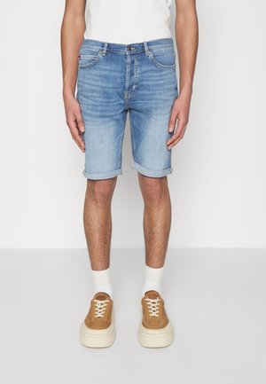 Man draagt lichtblauwe opgerolde spijkershorts, witte sokken en bruine sneakers, staand tegen een effen achtergrond.