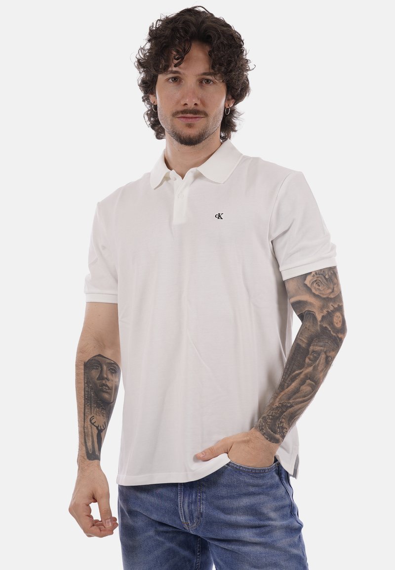 Homme aux cheveux bouclés et tatouages sur les deux bras, portant un polo blanc Calvin Klein et un jean bleu, une main dans la poche.