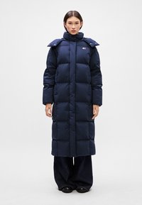 Dlhý námornícky prešívaný kabát s vysokým golierom, detailmi na patentky a kvadratickým strihom. Vyrobený z hladkého, lesklého materiálu.