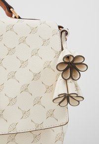 Borsa in pelle di avorio con texture cremosa, decorata con un motivo floreale dorato e tre dettagli a frange a forma di anello sui lati, manico marrone parzialmente visibile.