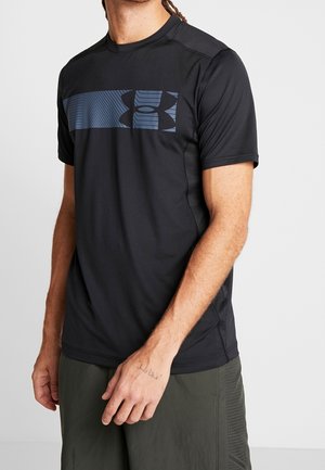 Mann trägt ein schwarzes kurzärmeliges Under Armour Shirt mit einem blauen Logostreifen über der Brust und dunkelgrüne Shorts.