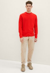 Rode gebreide trui met een ronde hals, gecombineerd met beige chinos en lichtbruine sneakers. Eenvoudig ontwerp, gladde textuur en getailleerde vorm.