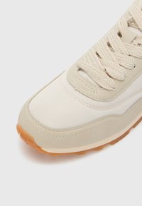 Beige sneaker with a smooth fabric upper, textured overlays, round toe, and rubber outsole featuring traction patterns. Laces are braided.