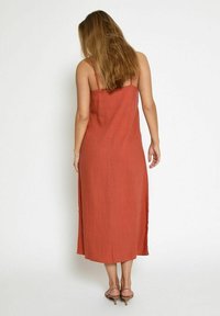 Robe mi-longue de couleur rouille en tissu léger, dotée de fines bretelles et de fentes sur les côtés pour faciliter les mouvements.