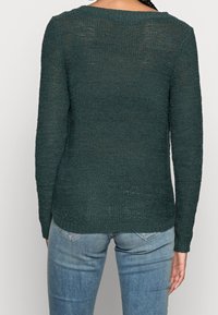 Donkergroen gebreide trui met lange mouwen, bootnek en structuurafwerking; gecombineerd met lichtblauwe jeans.