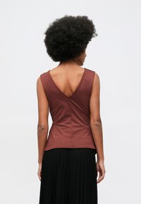 Γυναίκα με αφρο �χτένισμα, φορώντας αμάνικο καφέ τοπ με βαθύ V στην πλάτη και μαύρη μακριά πλισέ φούστα, που στέκεται μπροστά σε απλό φόντο.