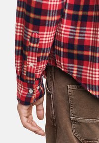 Czerwona i niebieska koszula w kratę z flaneli z białymi guzikami i mankietami zapinanymi na guziki, w połączeniu z brązowymi spodniami o teksturowanej powierzchni i z detalami bocznej kieszeni.