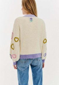 Krémový svetr s barevnými srdíčky v růžové, zelené a modré barvě. Obsahuje fialový žebrovaný lem a límec. Štítek je viditelný na zadní straně.