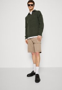 Pullover verde a maglia con zip a metà, pantaloni cargo beige, mocassini neri e calzini bianchi. Il modello è in piedi contro uno sfondo neutro.