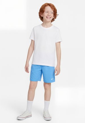 Chłopiec o rudych włosach, ubrany w białą koszulkę, jasnoniebieskie spodenki Nike, białe skarpetki i białe trampki, uśmiechający się i patrzący na bok.