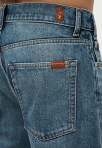 Πίσω τσέπη από τζιν με καφέ ετικέτα μάρκας και μικρό δερμάτινο έμπλαστρο στη μέση, φορεμένη από ένα άτομο.