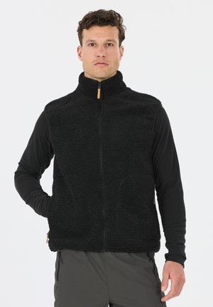 Man staat recht vooruit kijkend, draagt een zwart fleece vest met rits over een shirt met lange mouwen, met een hand in de vestzak tegen een egale achtergrond.