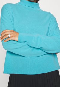 Pull à col roulé turquoise en tricot doux. Il présente une coupe décontractée et des accents côtelés au niveau du col et des poignets.