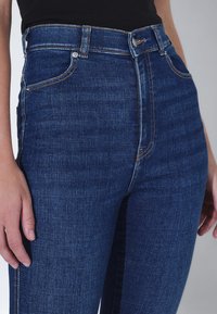 Jeans de mezclilla azul de talle alto con una silueta ajustada, bolsillos frontales y traseros, y costuras en color cobre.