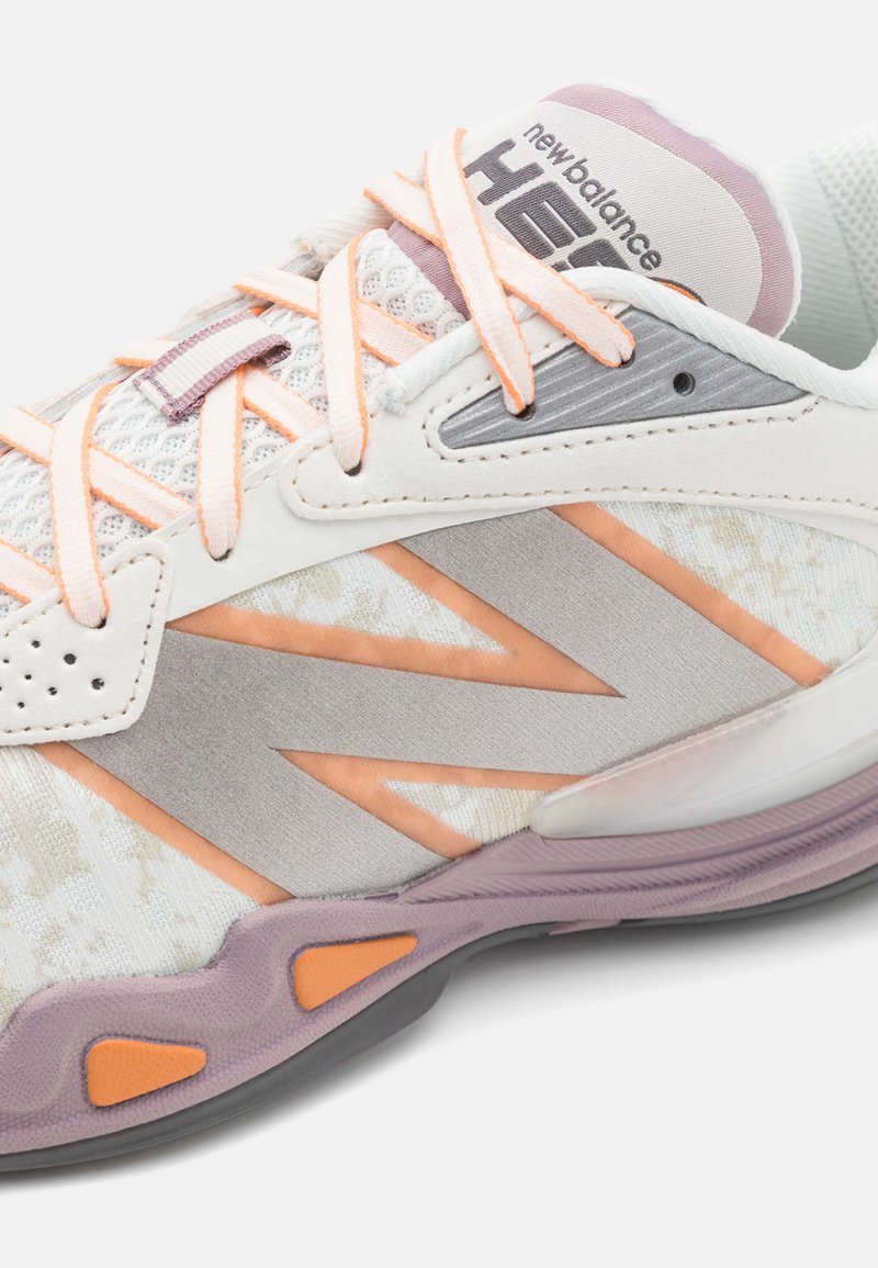 Zapatilla New Balance blanca y morada clara con acentos en melocotón, lengüeta de malla y cordones naranja claro en vista en primer plano.