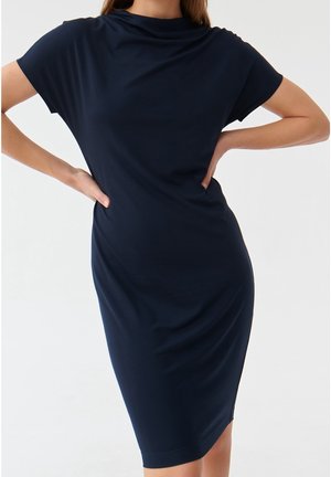 Femme portant une robe midi bleu marine ajustée à manches courtes, les mains sur les hanches, sur un fond clair uni.